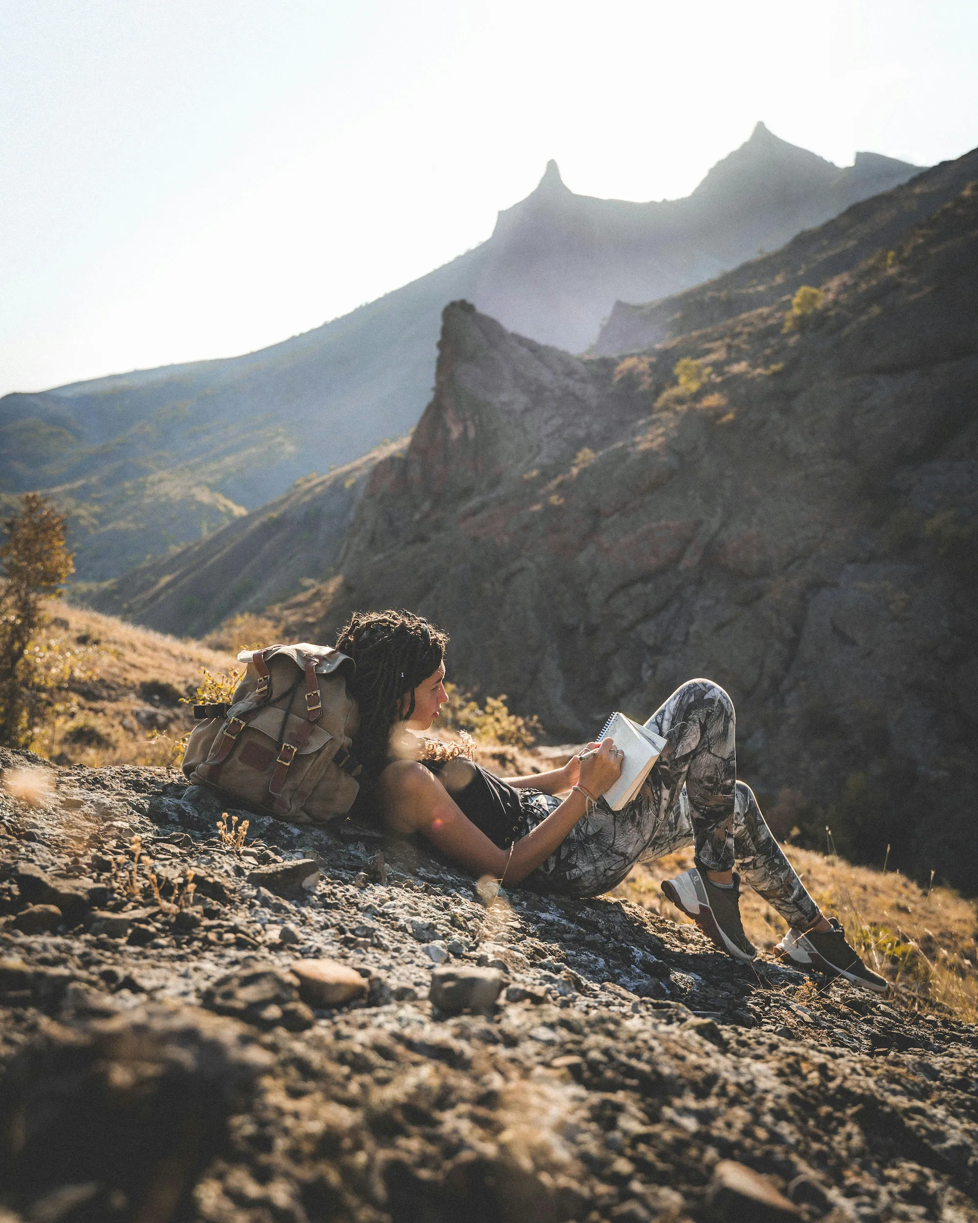 Traveler lying on a rocky hillside writing in a notebook, with mountains behind